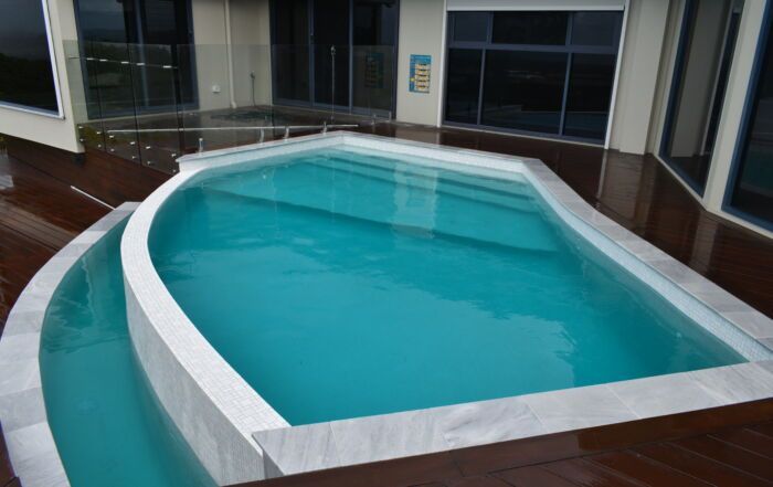 Curved Indoor Pool with Turquoise Water and White Coping Beside Wooden Deck — Mad Pool Interiors and Renovations in Baringa, QLD