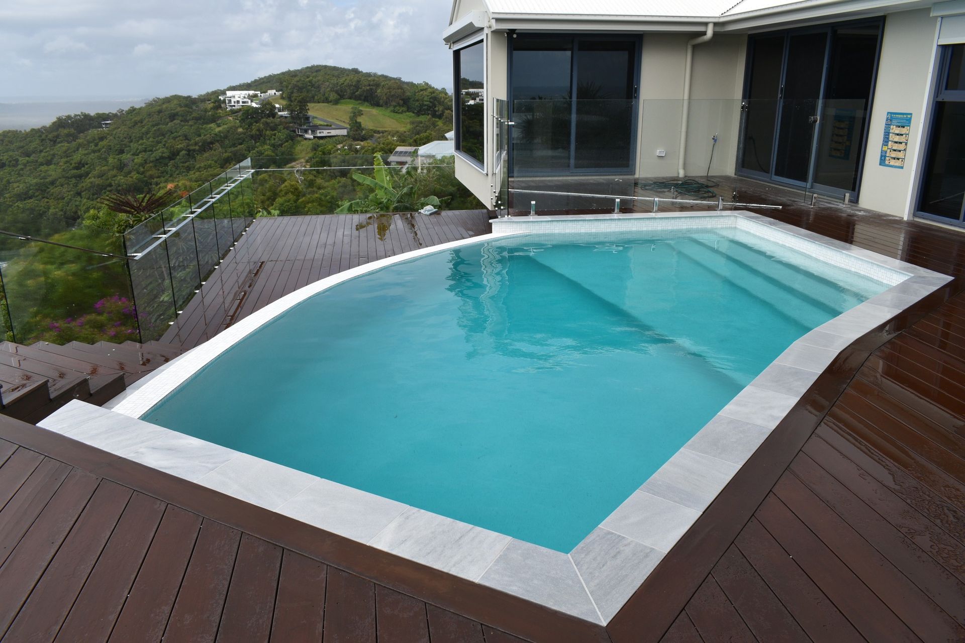 A Turquoise Swimming Pool With a Curved Edge on a Dark Wooden Deck — Mad Pool Interiors and Renovations in Baringa, QLD