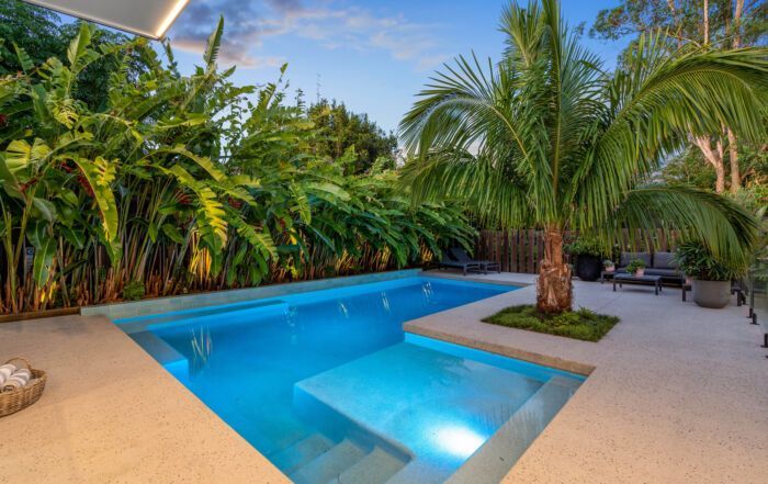 Tropical Backyard Pool with Blue Water, White Deck, Palm Trees, and Lush Greenery at Sunset — Mad Pool Interiors and Renovations in Baringa, QLD