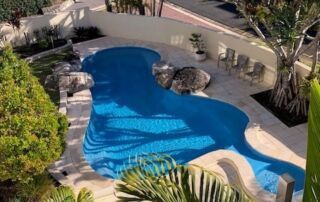 Backyard Swimming Pool with Curved Blue Water, Stone Deck, and Surrounding Palm Trees — Mad Pool Interiors and Renovations in Baringa, QLD