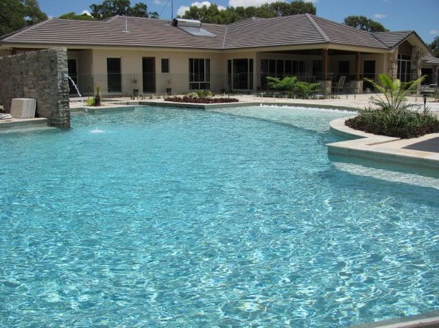 A Large Blue Swimming Pool With a House in the Background on a Sunny Day — Mad Pool Interiors and Renovations in Baringa, QLD