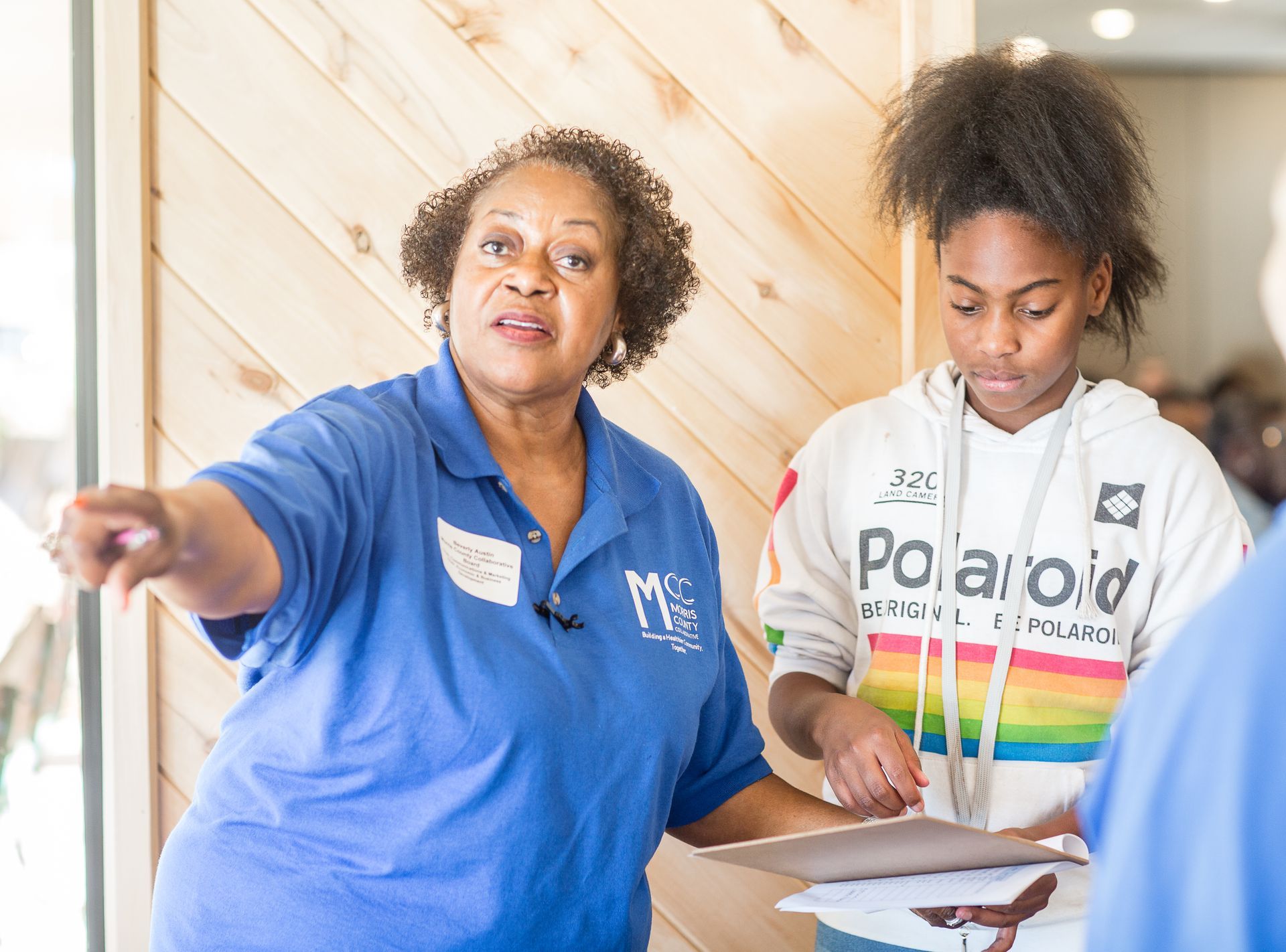 A woman in a blue shirt is talking to a girl in a polaroid hoodie.