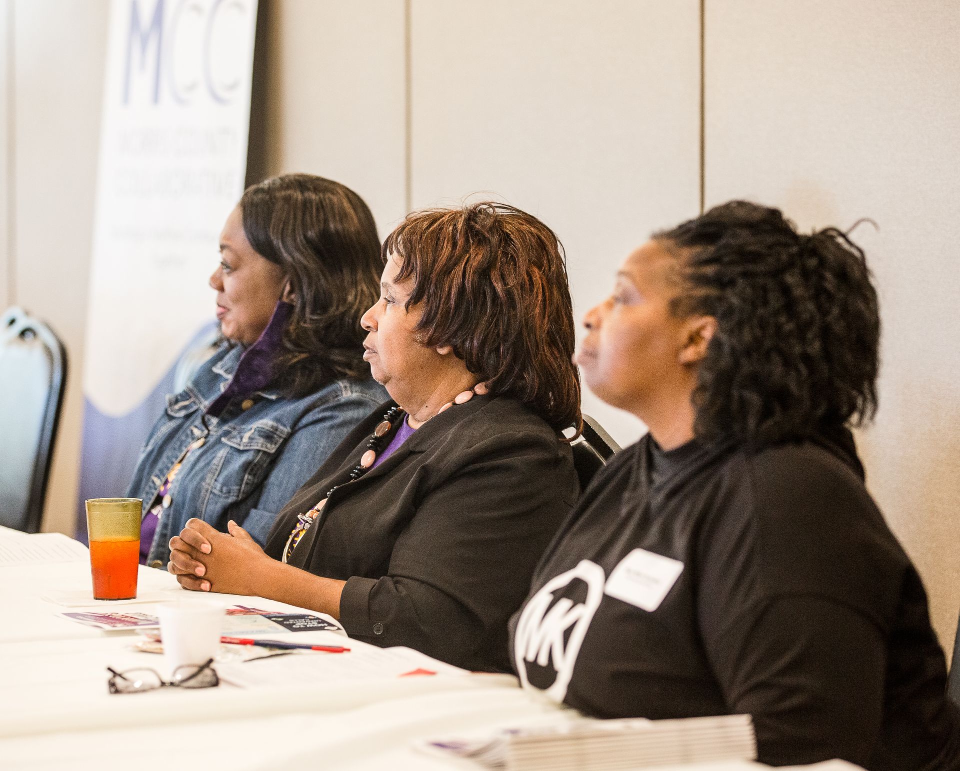 Three women are sitting at a table in front of a mcc banner