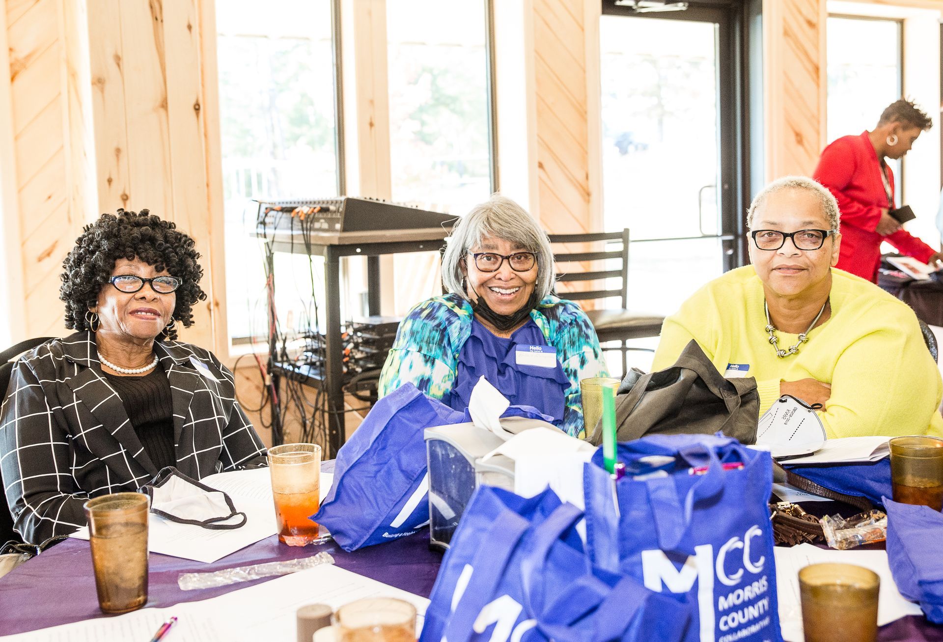 Three women are sitting at a table with a bag that says mcc on it.