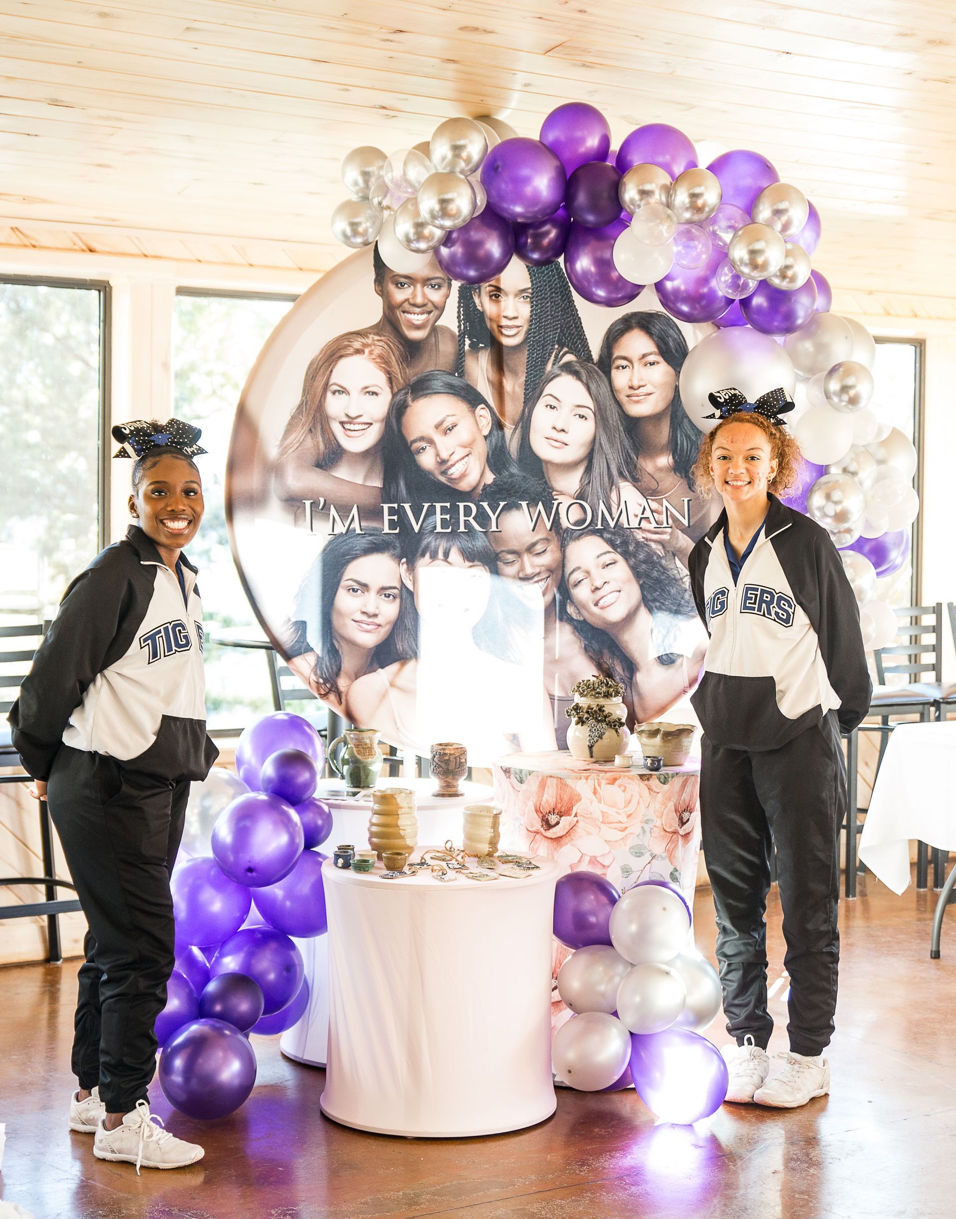 Two women are standing in front of a table decorated with purple and silver balloons.
