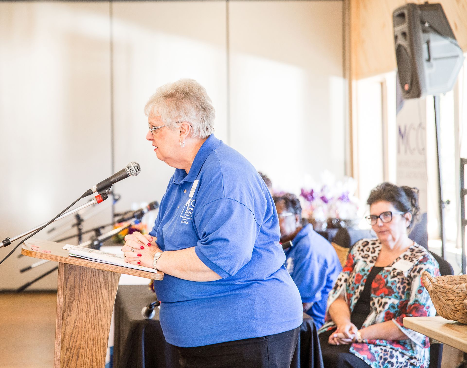 A woman in a blue shirt is standing at a podium speaking into a microphone.