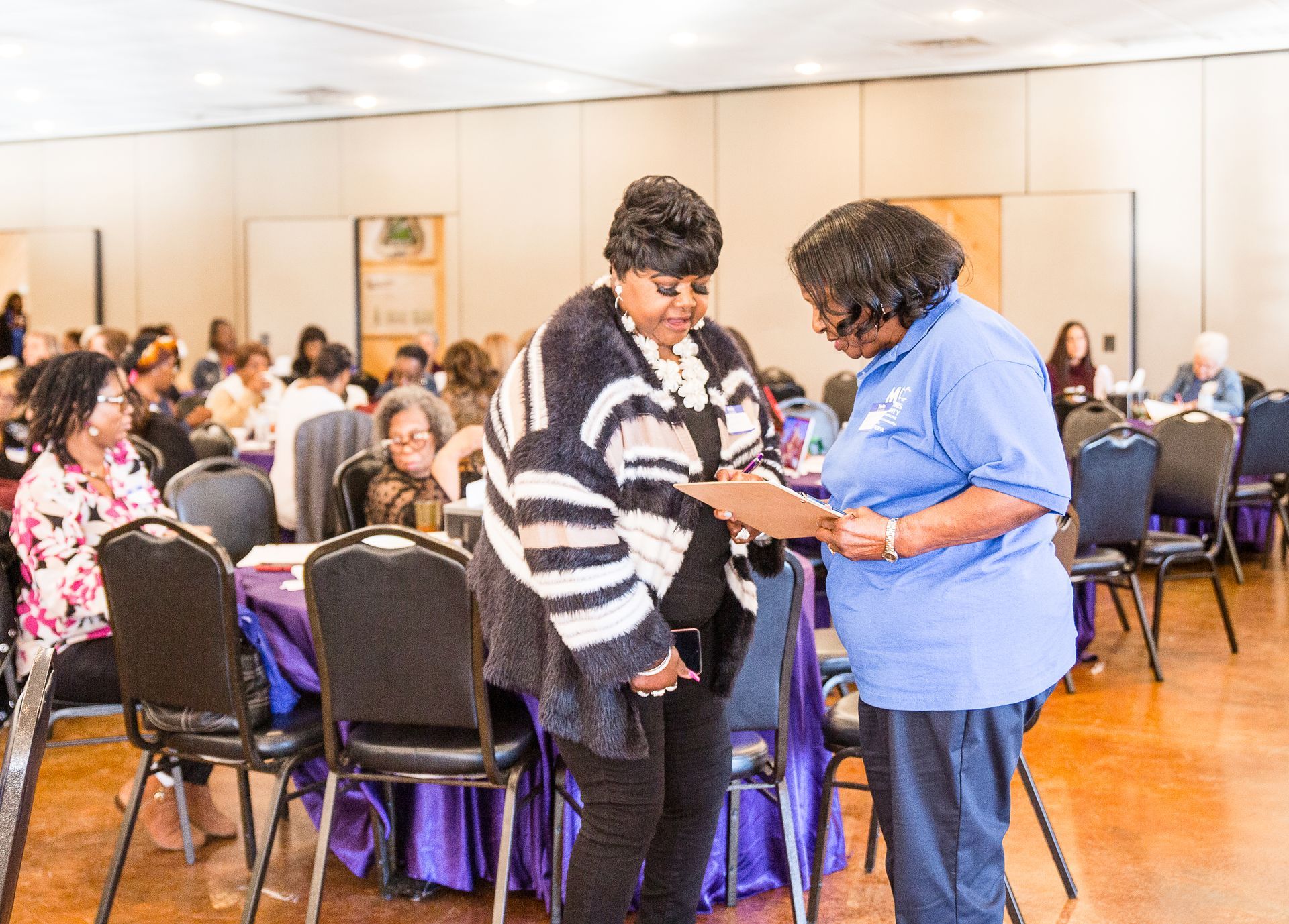 Two women are standing next to each other in a room with people sitting at tables.