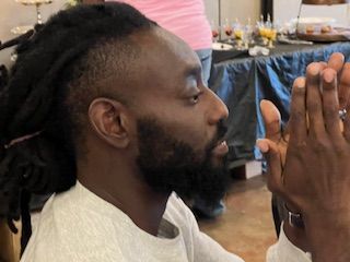 A man with dreadlocks and a beard is sitting at a table with his hands together.