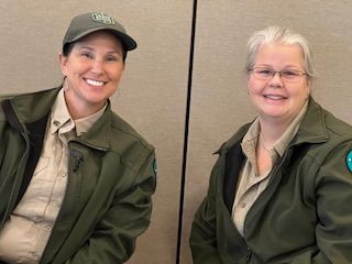 Two women wearing green jackets and hats are sitting next to each other and smiling.
