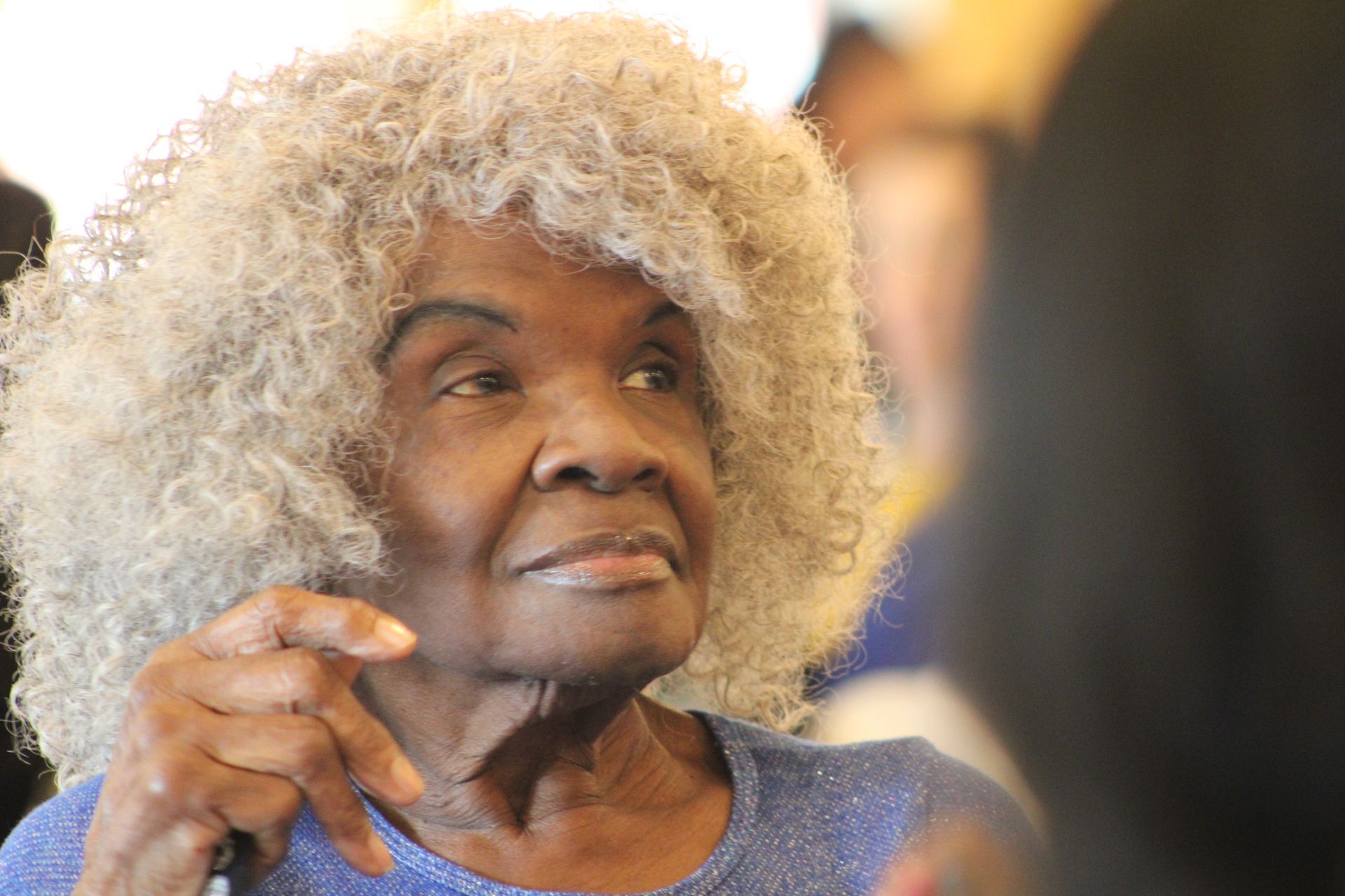 An elderly woman with gray curly hair is wearing a blue shirt.