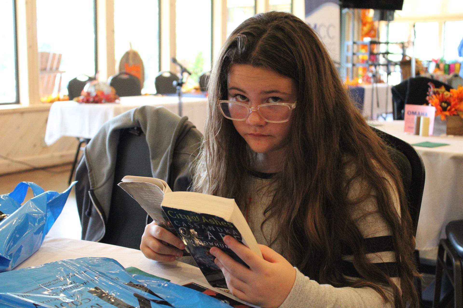 A young girl wearing glasses is reading a book at a table.
