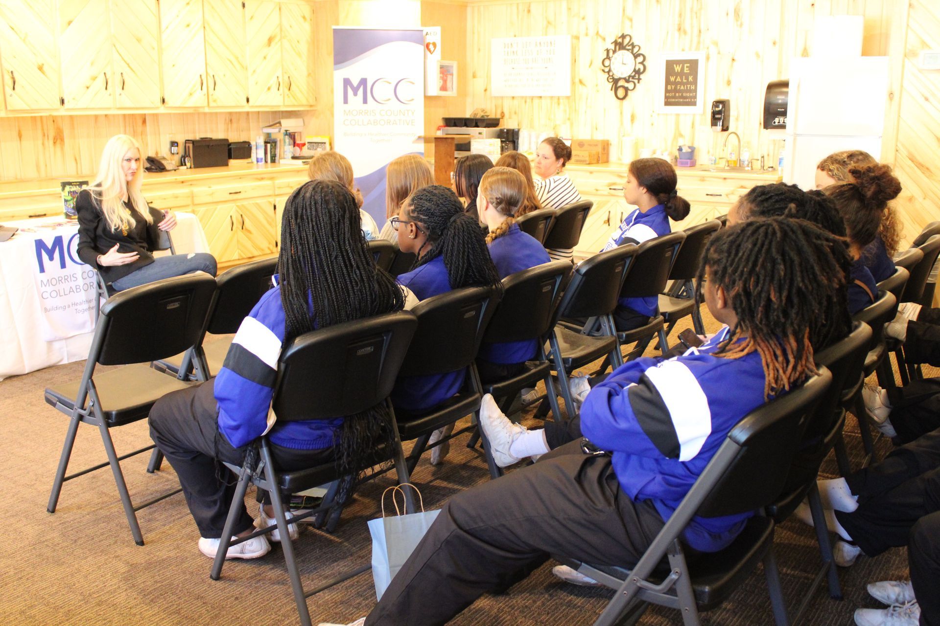 A group of people are sitting in chairs in front of a mcc sign