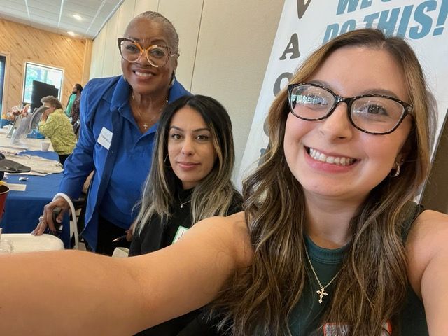 Three women are posing for a selfie in front of a sign that says we do this.