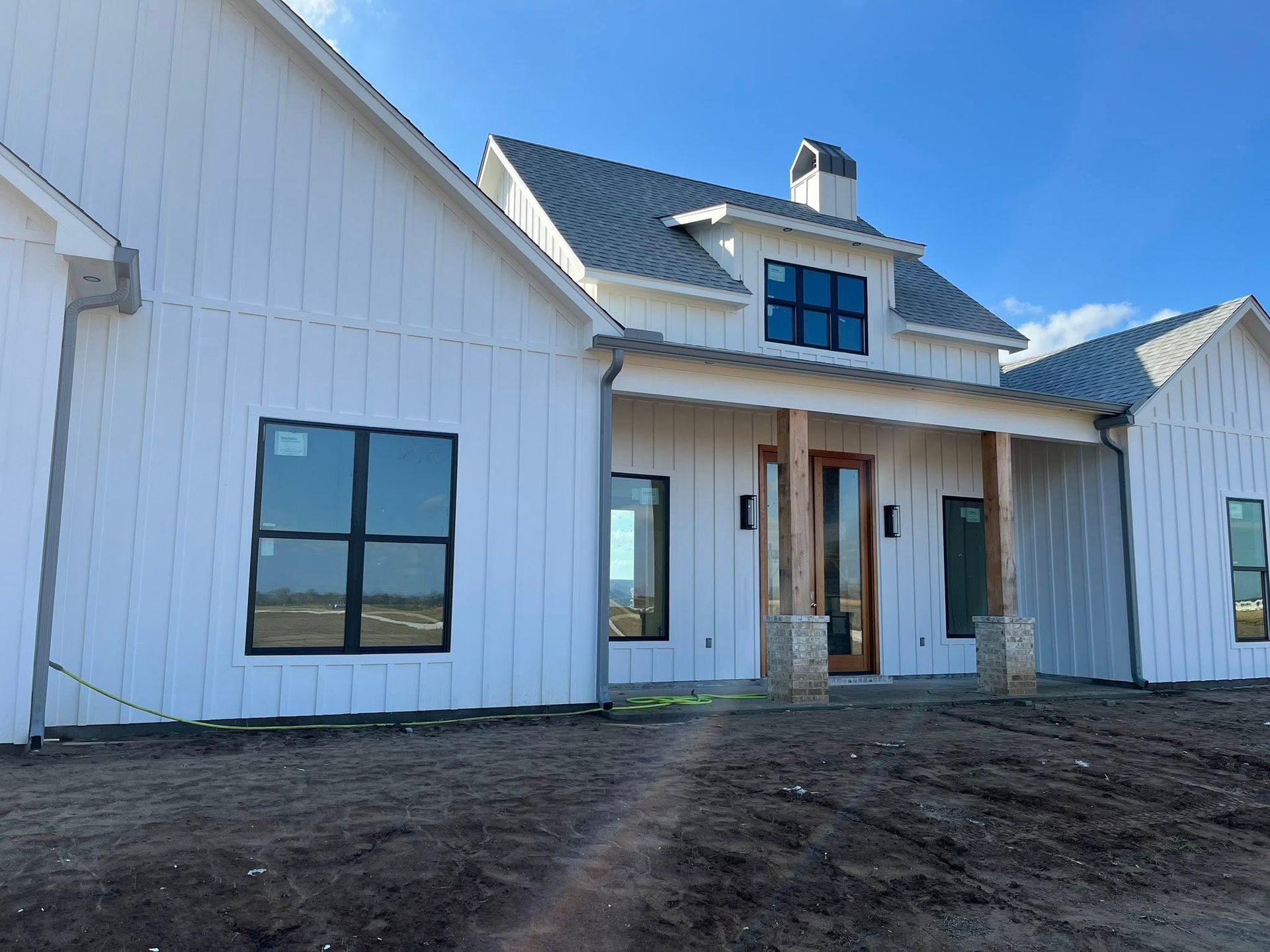 White farmhouse exterior with black-trim windows, covered porch, and dirt front yard under blue sky