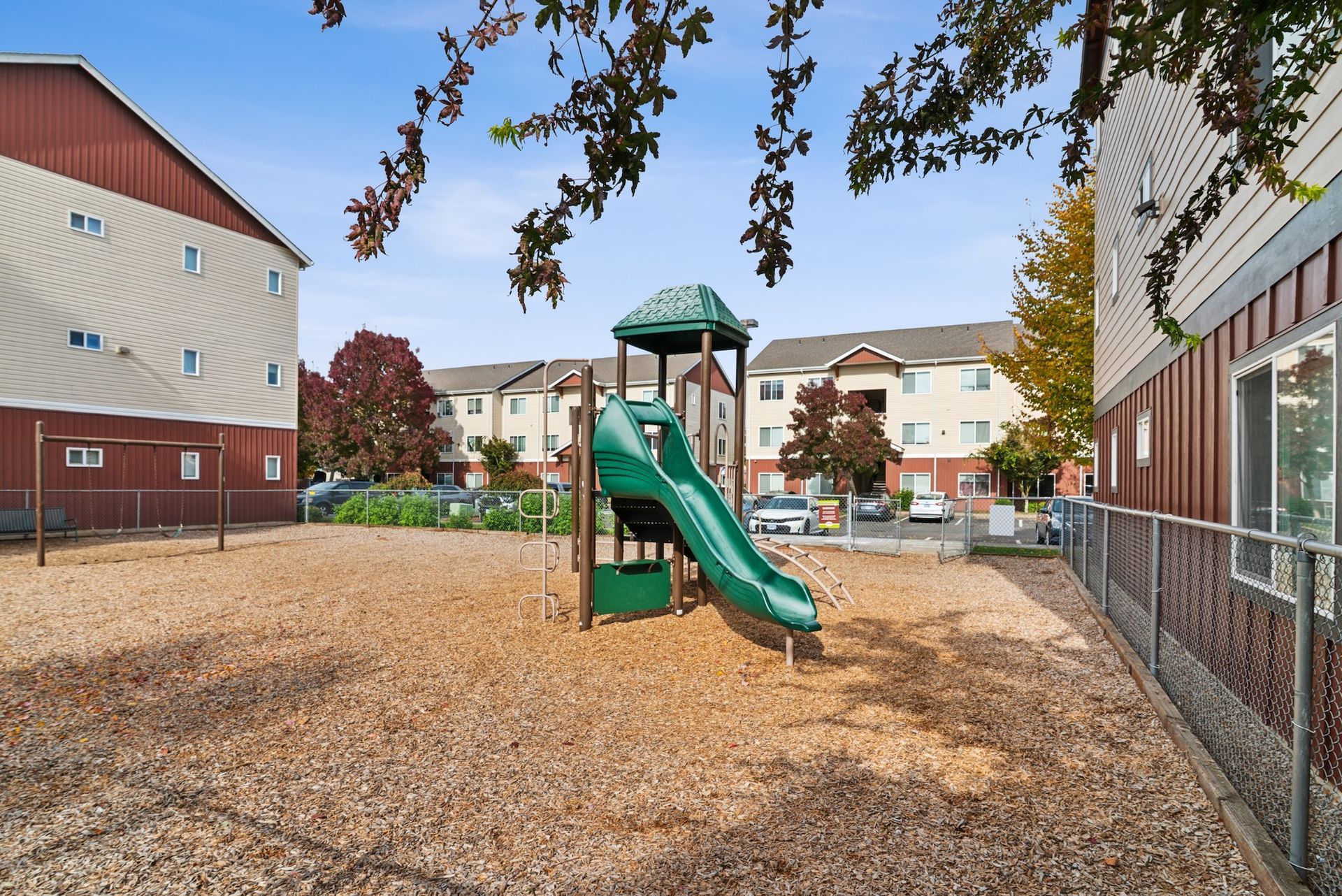 Playground with a green slide, surrounded by wood chips, in front of apartment buildings.