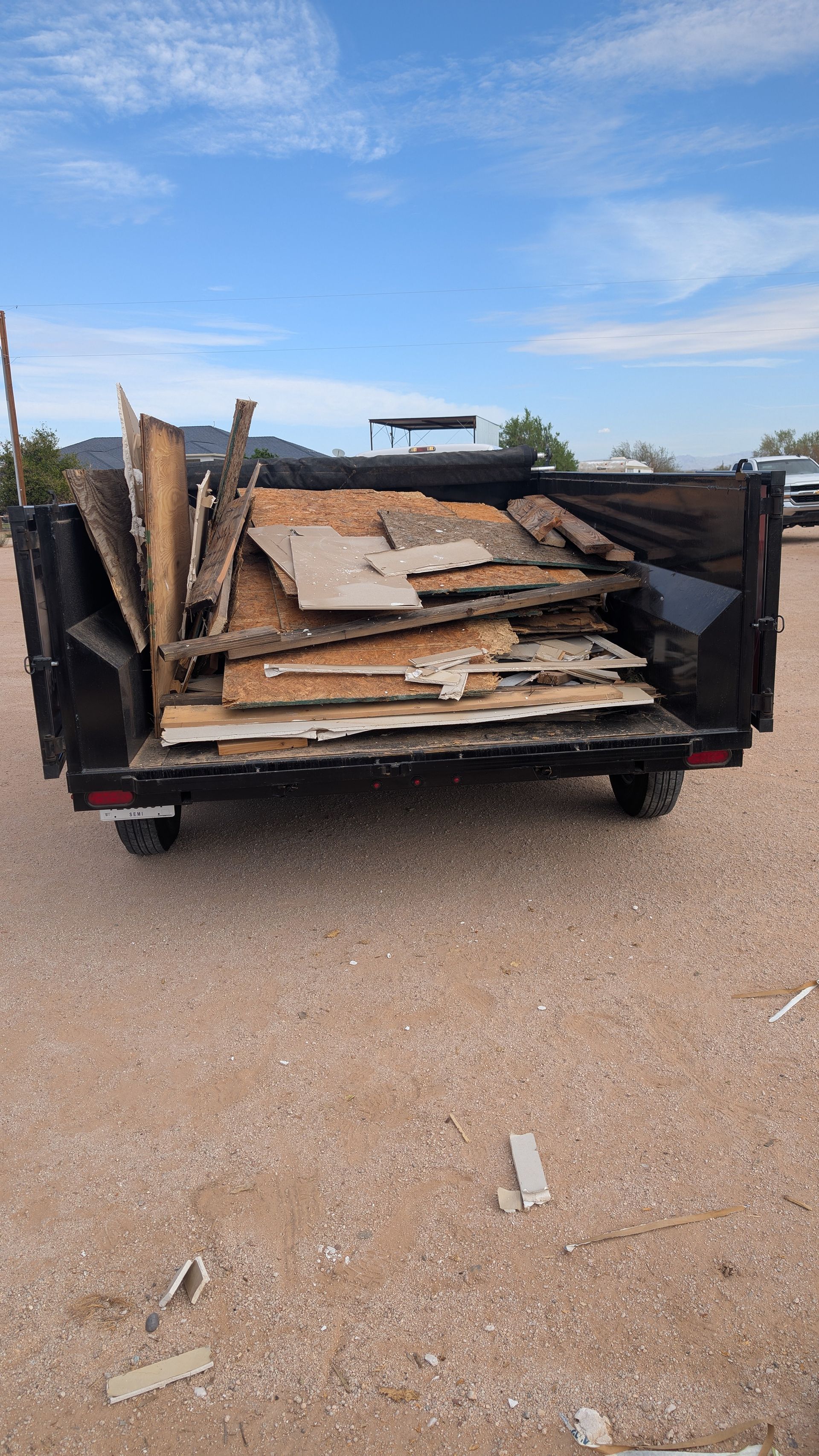 A trailer filled with old wood, debris, and scrap, parked on a gravel surface under a blue sky.
