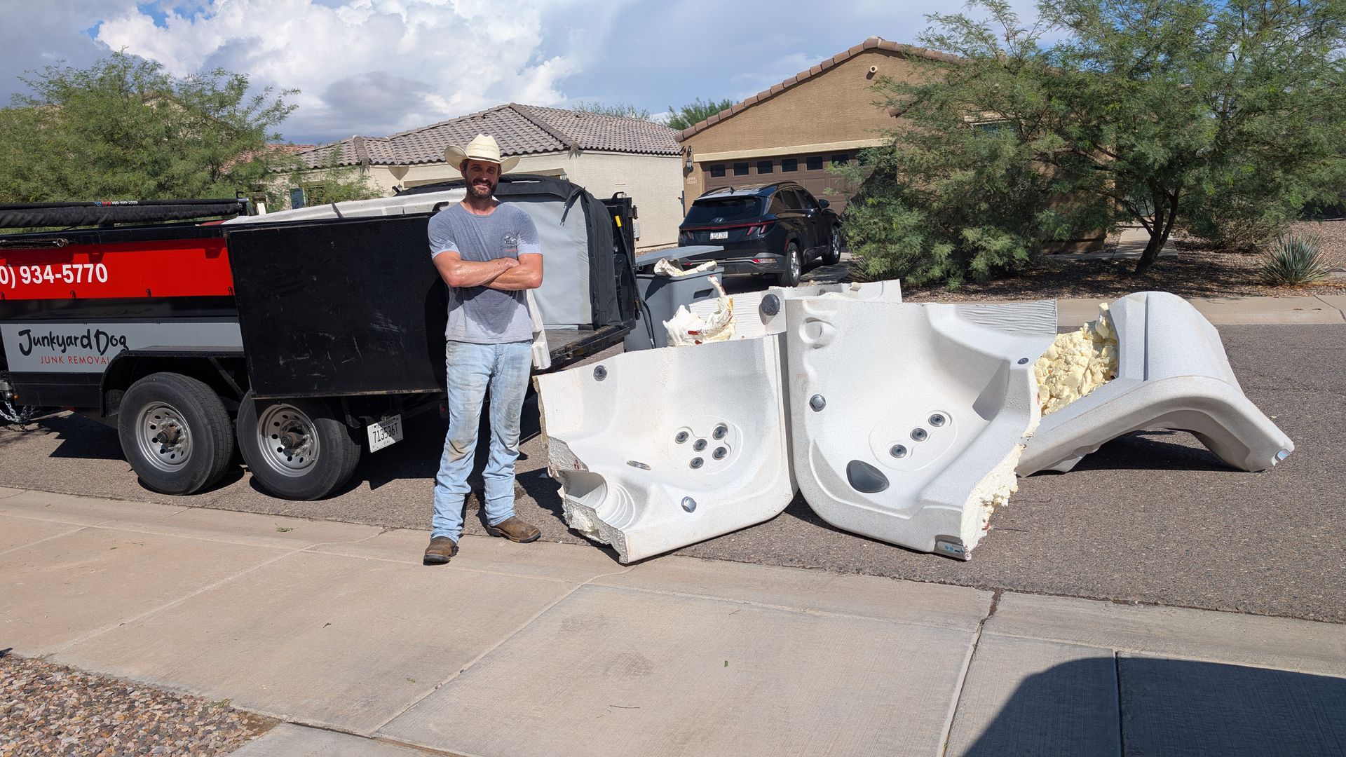 Man stands with arms crossed next to a broken hot tub and a junk removal truck on a street.