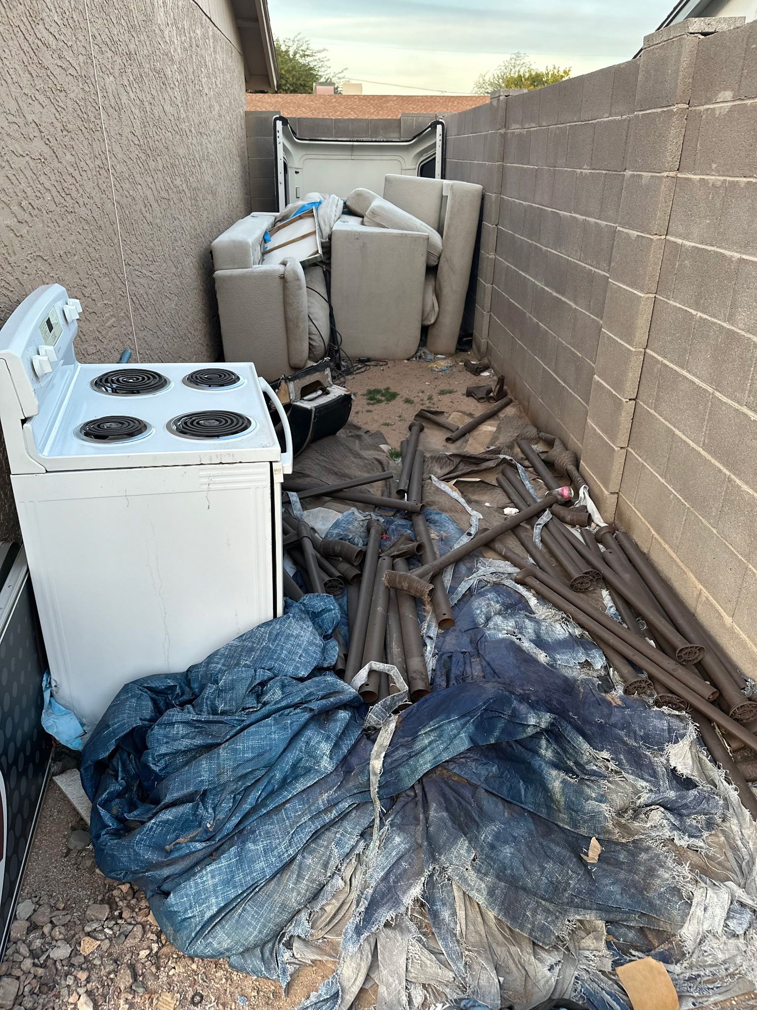 Backyard alleyway with discarded stove, debris, and trash bags against a cinder block wall.