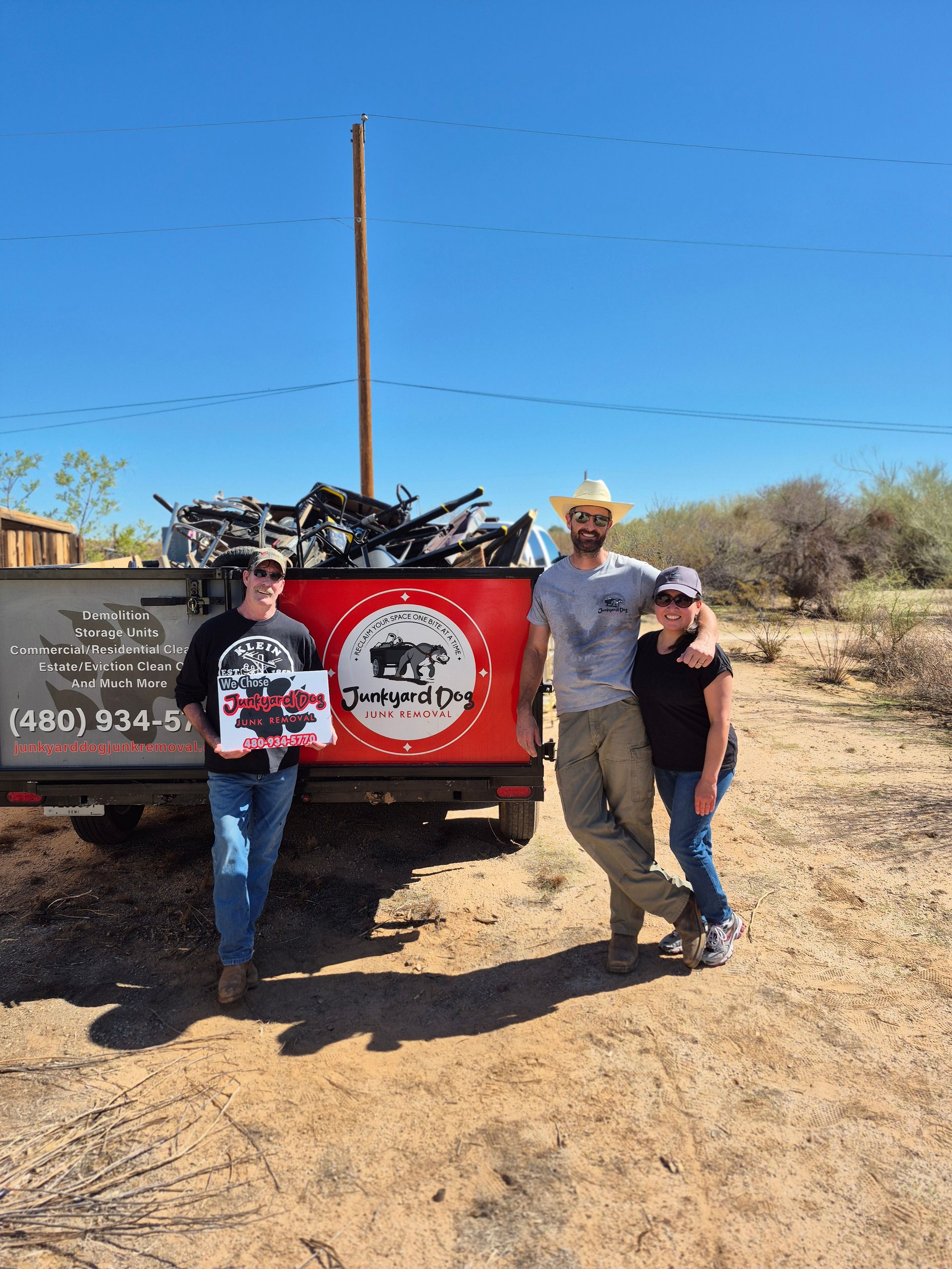 Three people pose in front of a truck loaded with scrap metal under a blue sky; one holds a sign for a junk removal service.