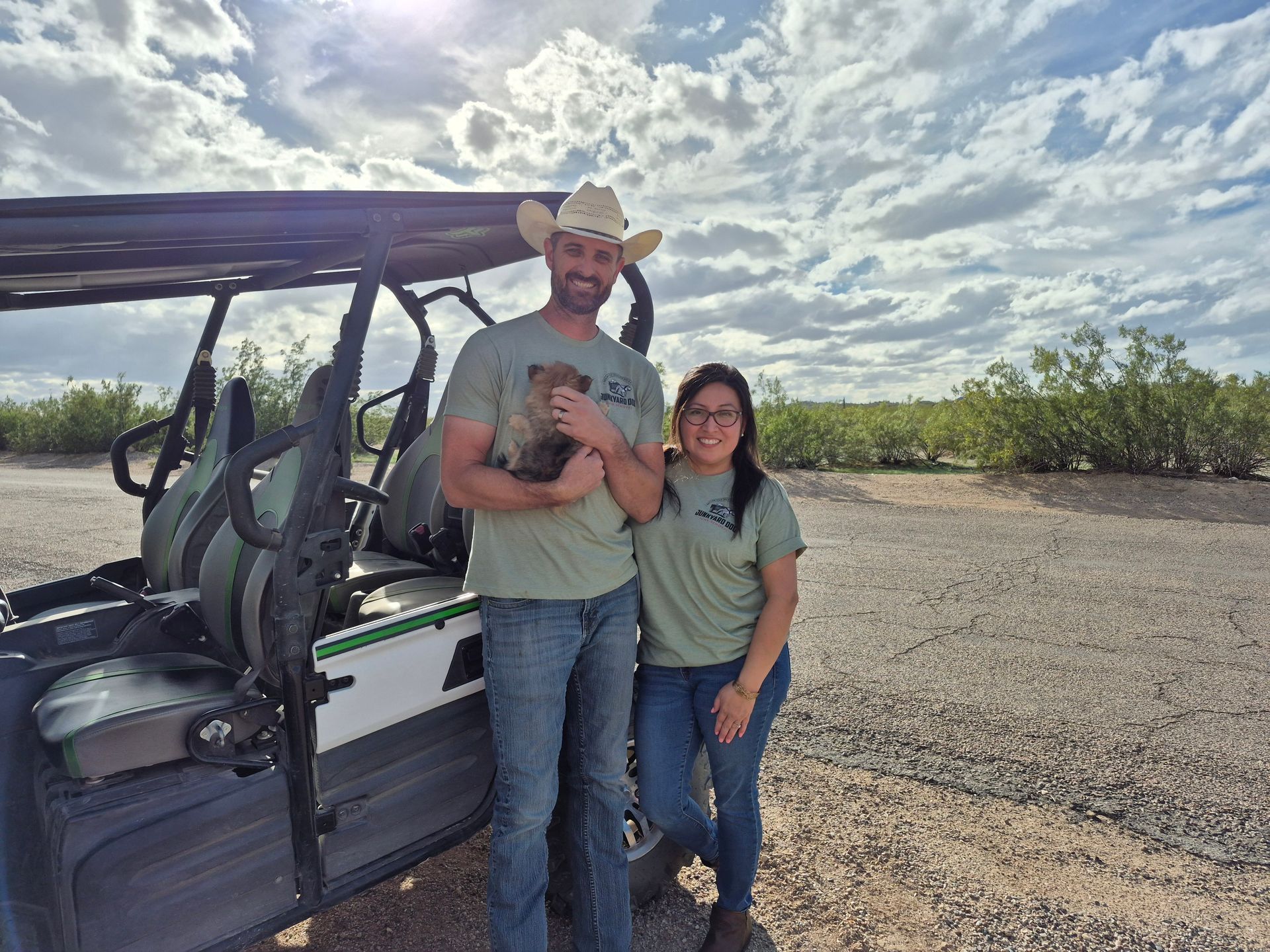 Man in cowboy hat holds small animal next to a woman; they stand by a golf cart outdoors.