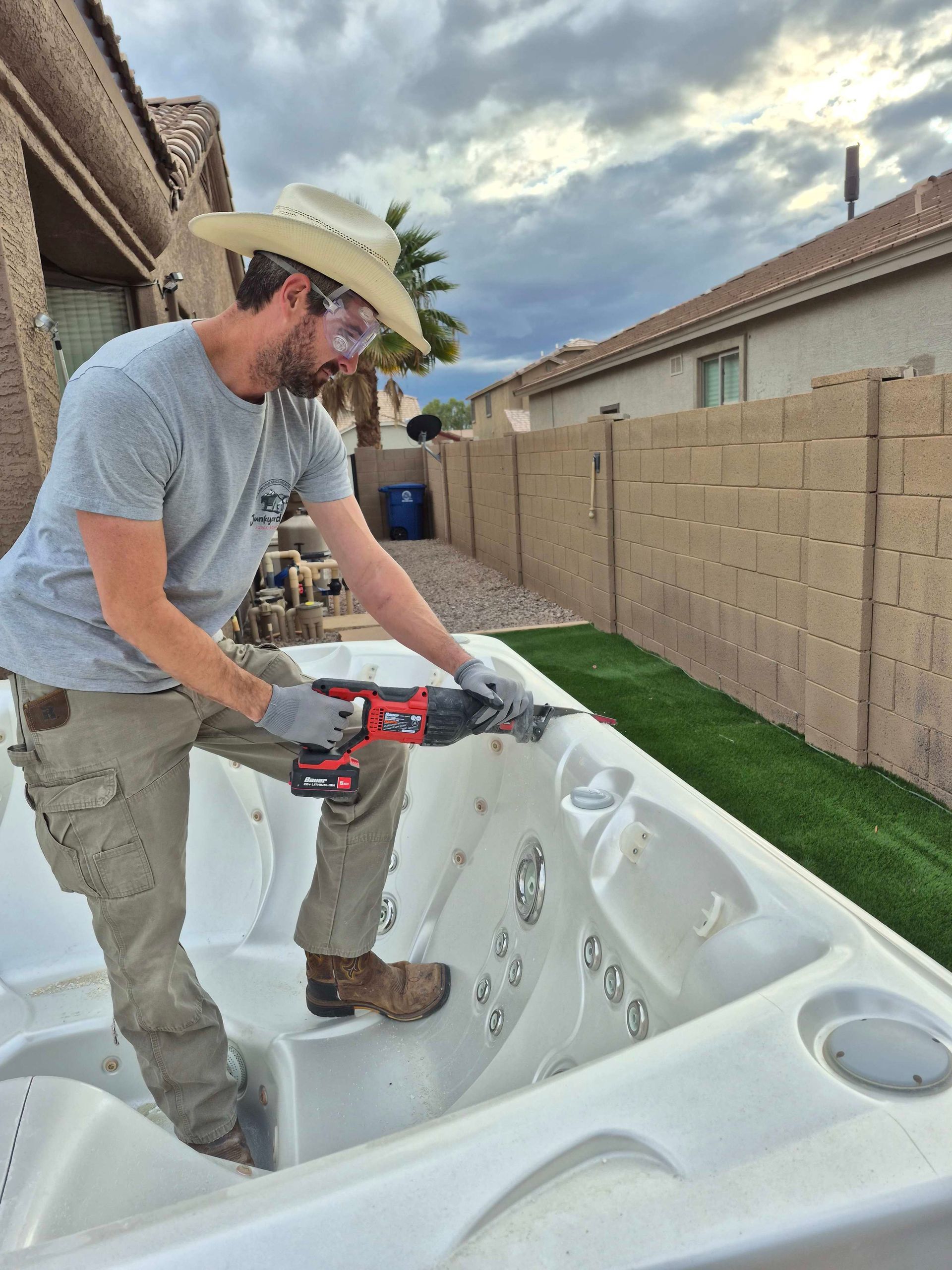 Man in cowboy hat uses a power saw on a white hot tub, outdoors next to a fence.
