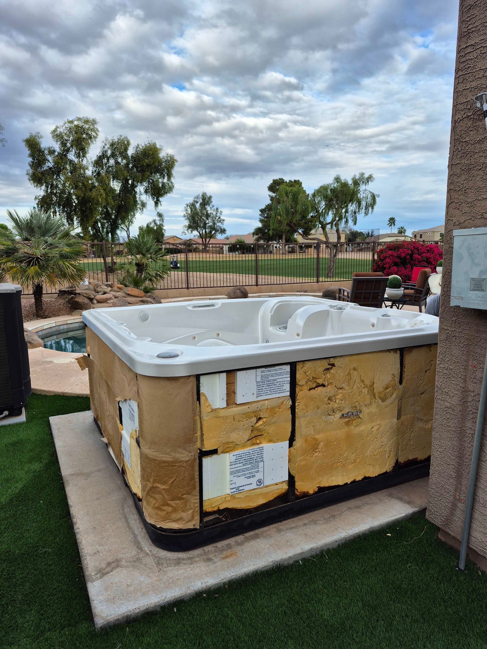 Hot tub with exposed insulation sits on concrete near a house, pool, and green lawn under a cloudy sky.