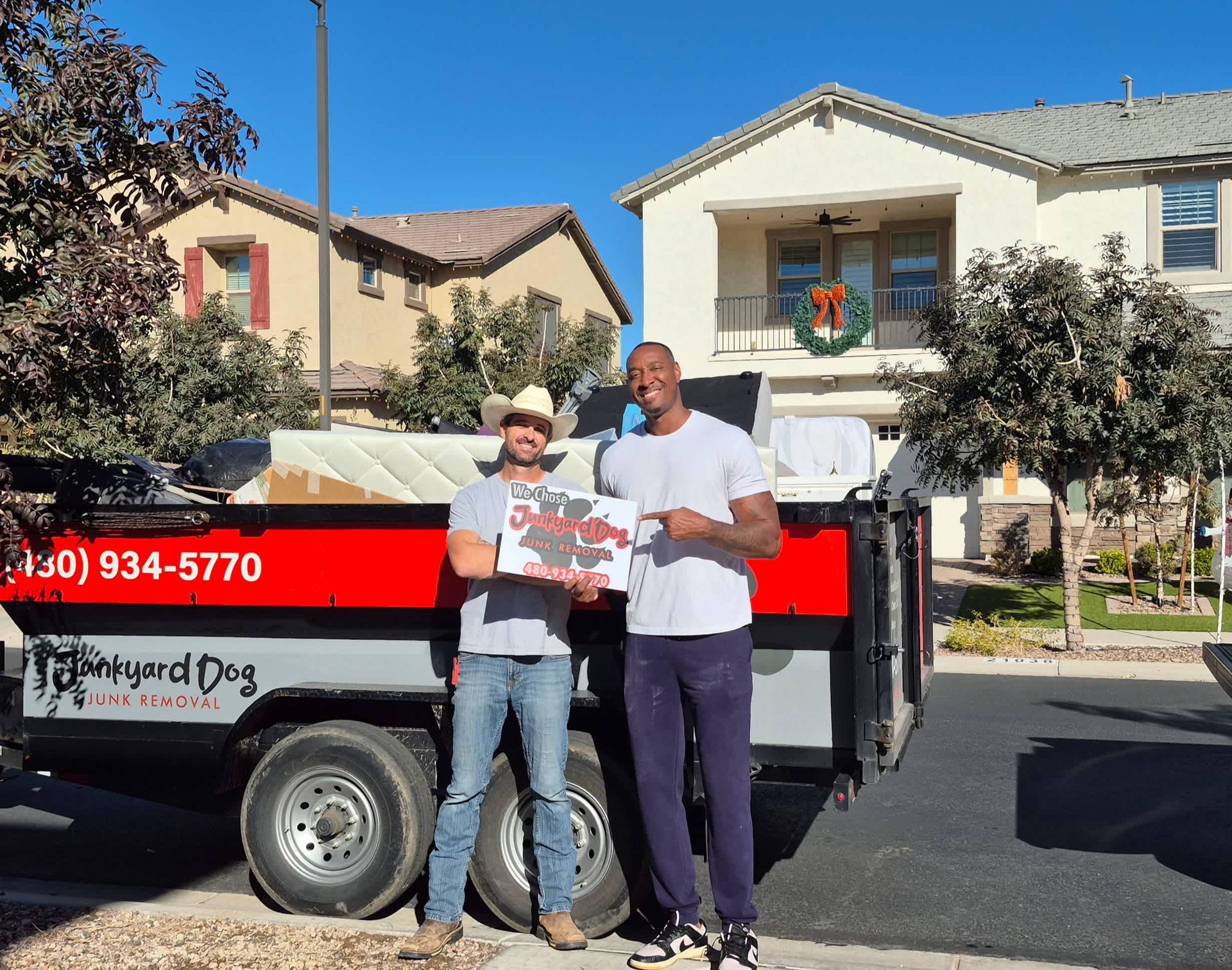Two men standing in front of a trailer filled with debris and holding a sign; in front of suburban homes on a sunny day.
