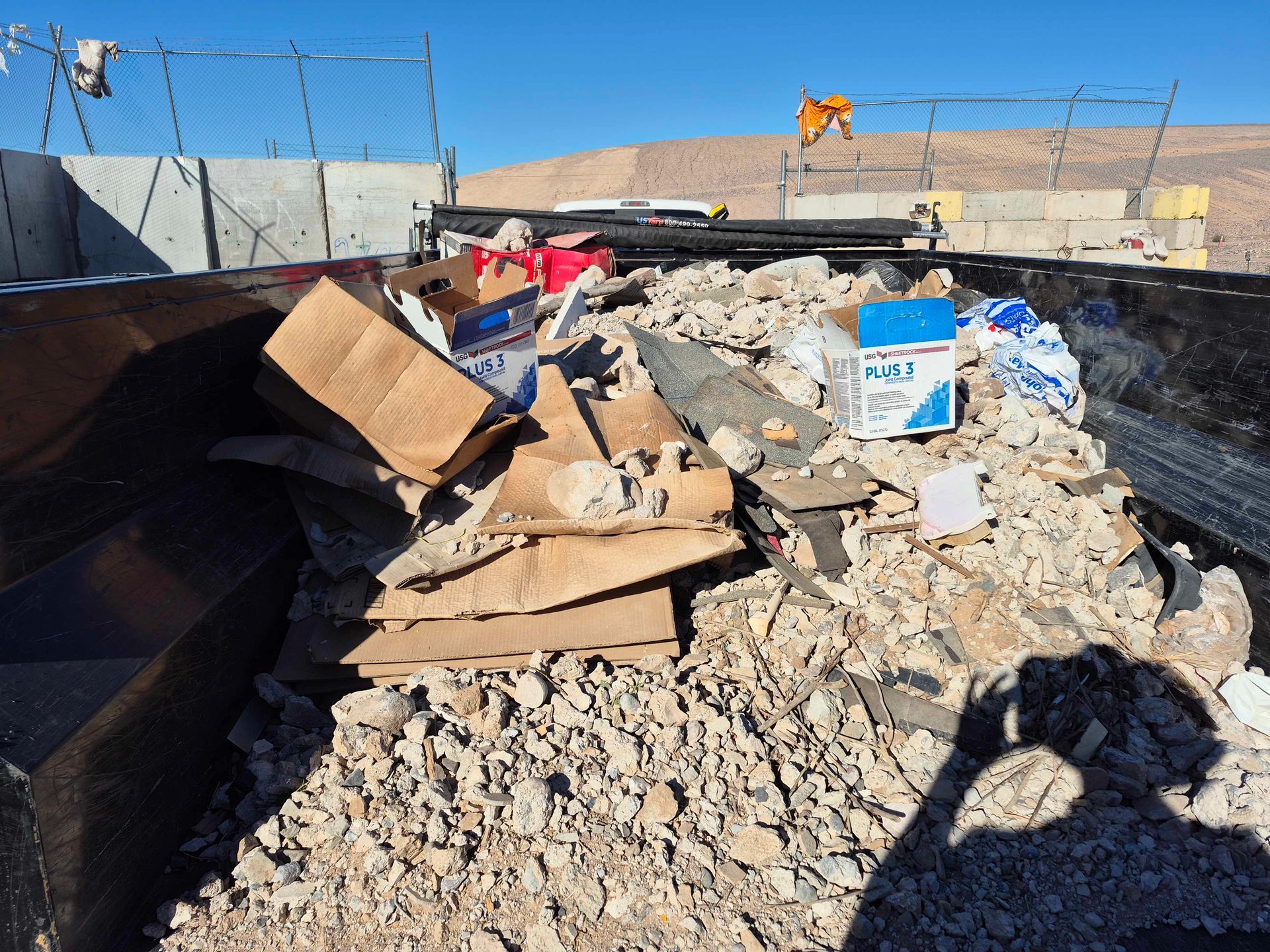 Dumpster filled with debris, including cardboard and broken concrete, against a fence and blue sky.