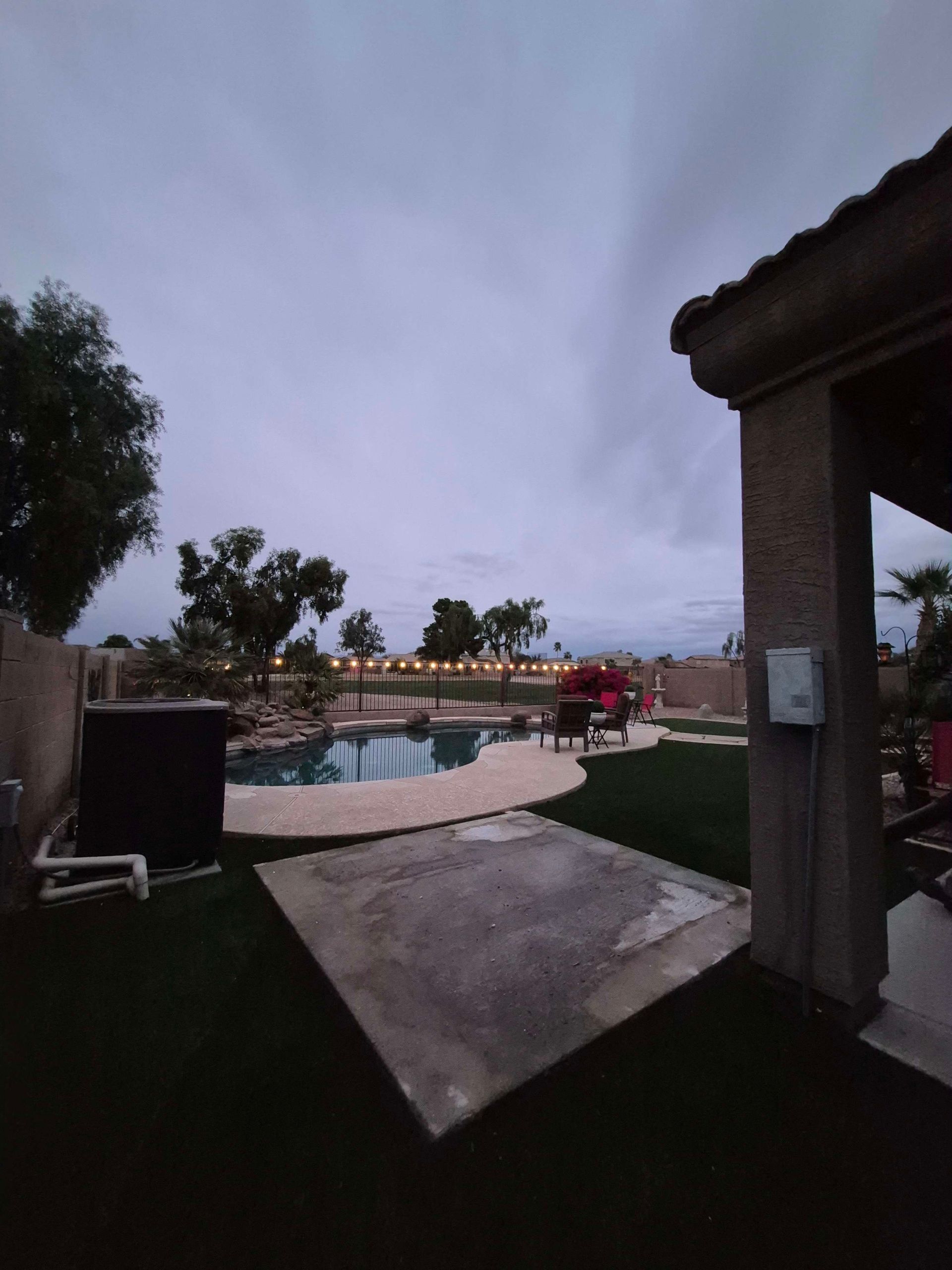 Backyard with pool, overcast sky. Stone patio, green grass, and trees. House structure on right.