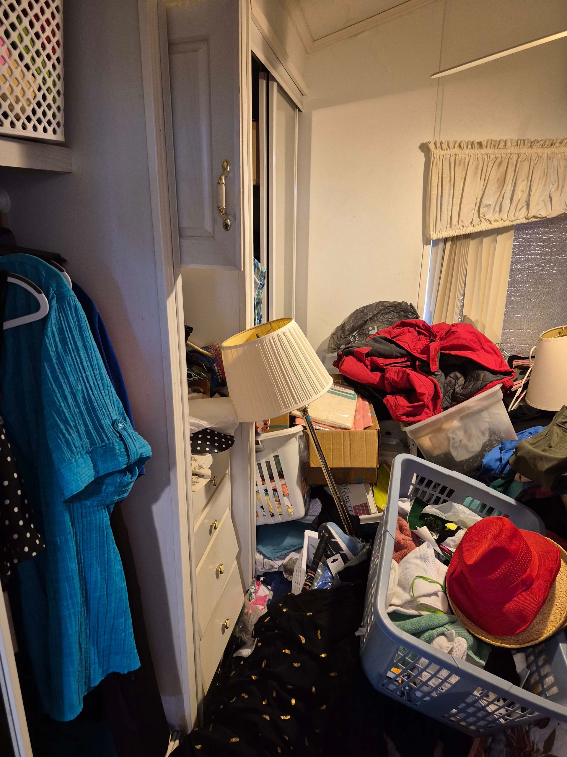 Messy bedroom: clothes scattered, baskets overflowing, lamp fallen. Wardrobe on left, window on right.