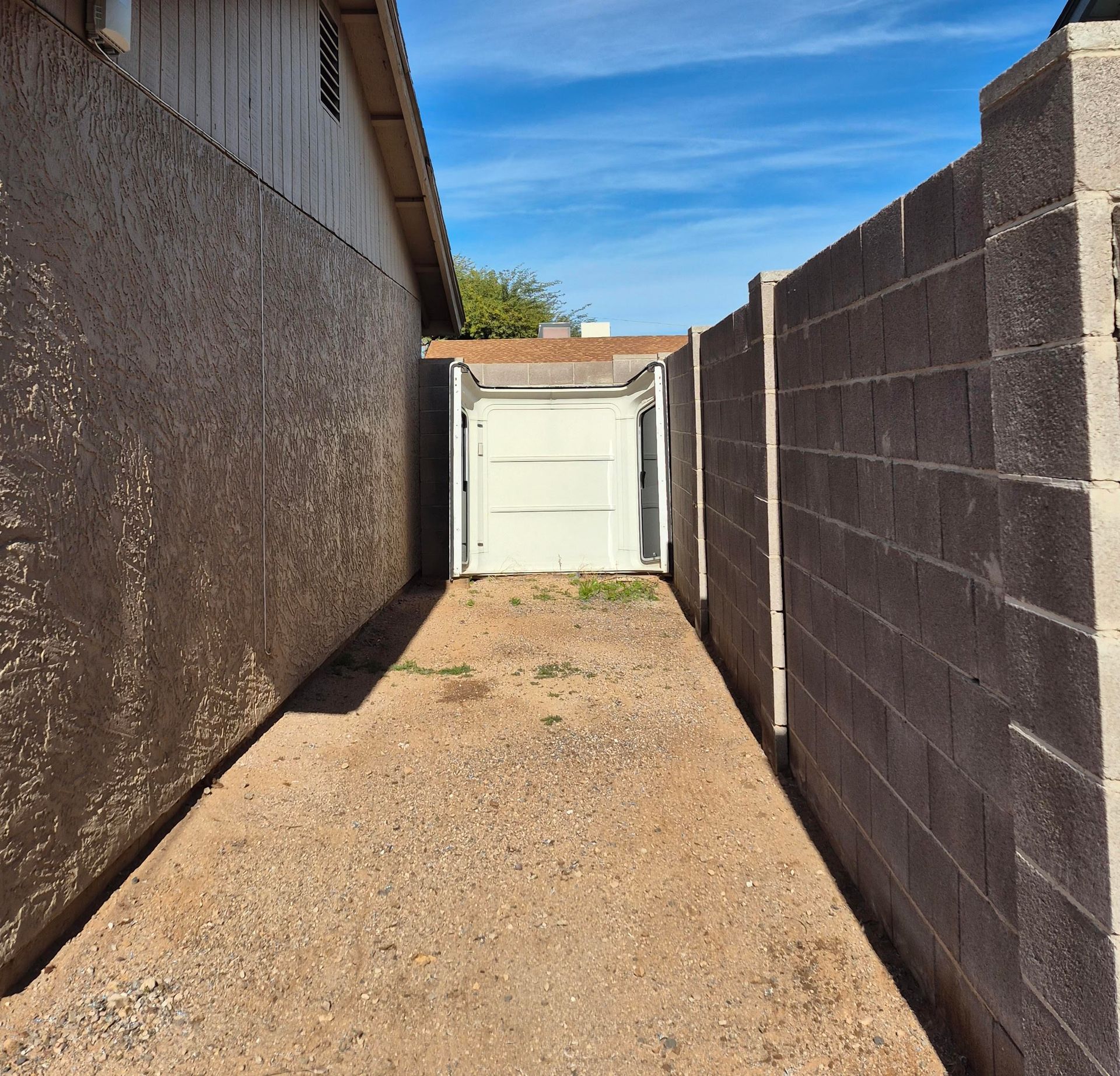 A narrow alleyway with a gravel ground, flanked by concrete walls, leads to a white garage door.