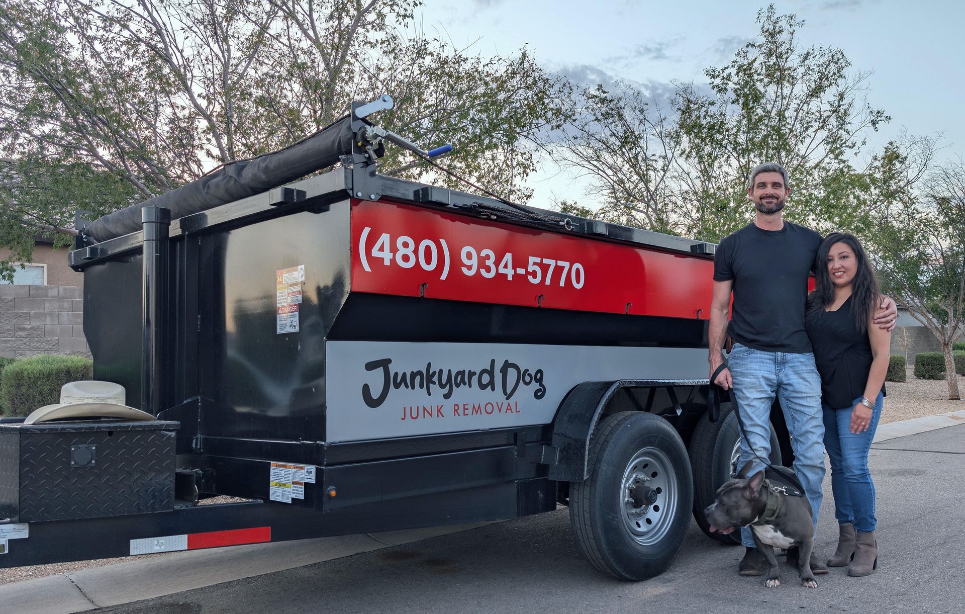 Man and woman pose with a blue nose pit bull in front of a dumpster trailer with 