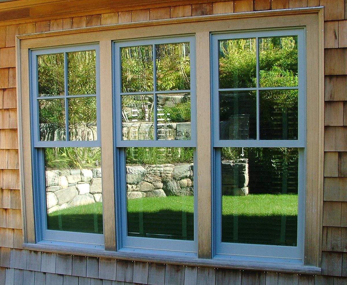 Three vertical windows with blue trim, reflecting a green yard, set in a wood-shingled wall.