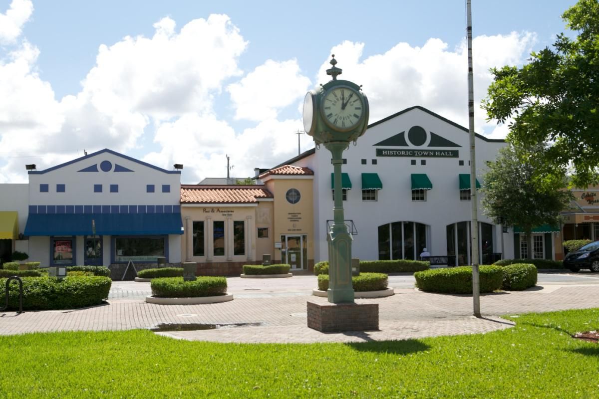 Clock tower in a town square, with shops and buildings in the background. Green grass and blue sky.