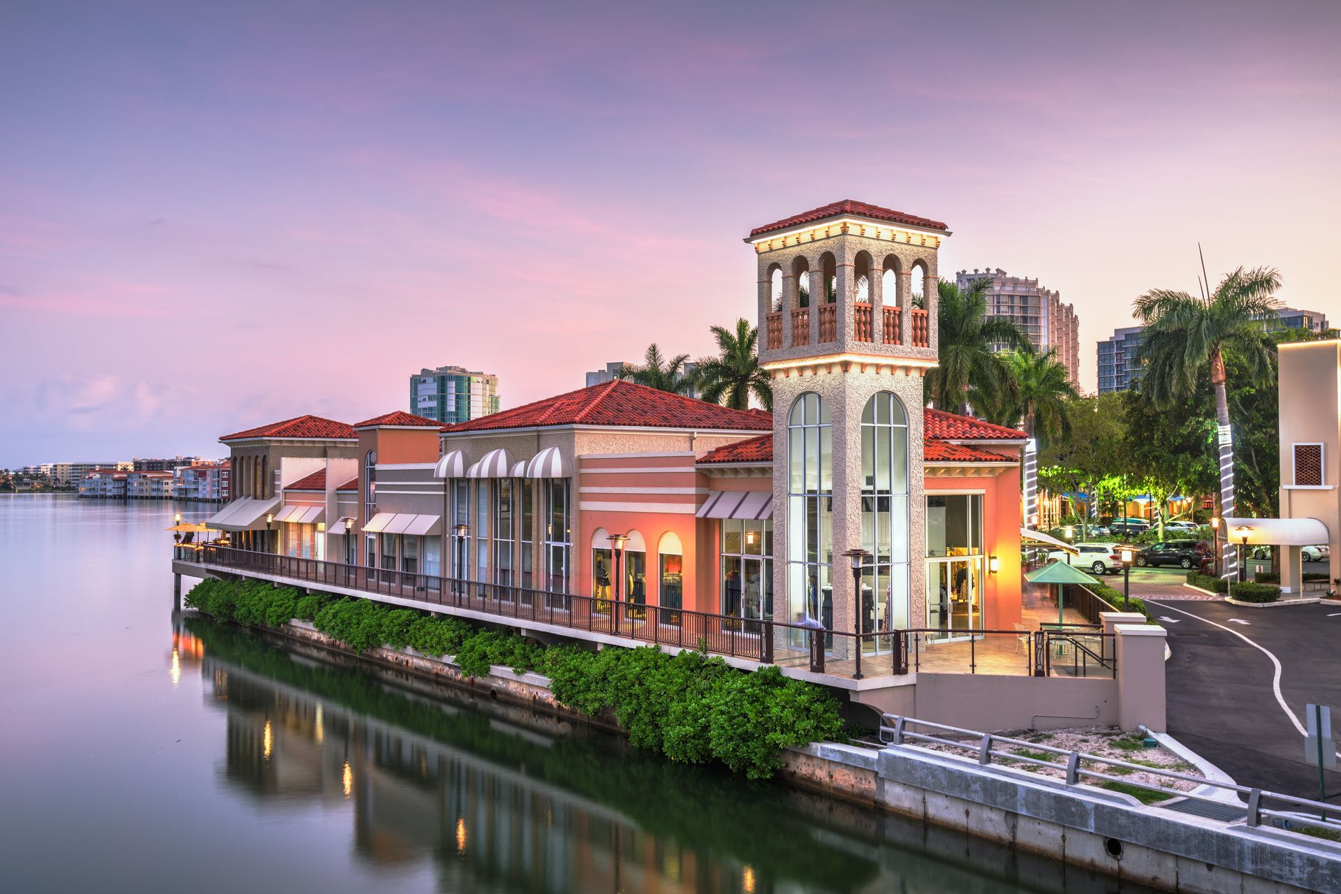 Waterfront buildings with a tower under a dusky sky with palm trees.