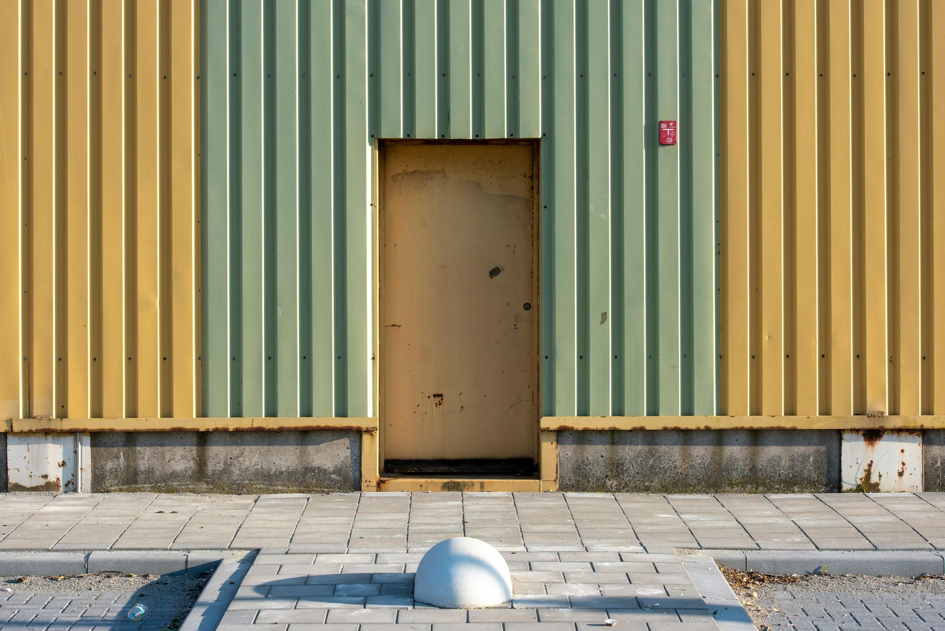 Yellow and green corrugated metal wall with a central, closed door; a concrete walkway in front.