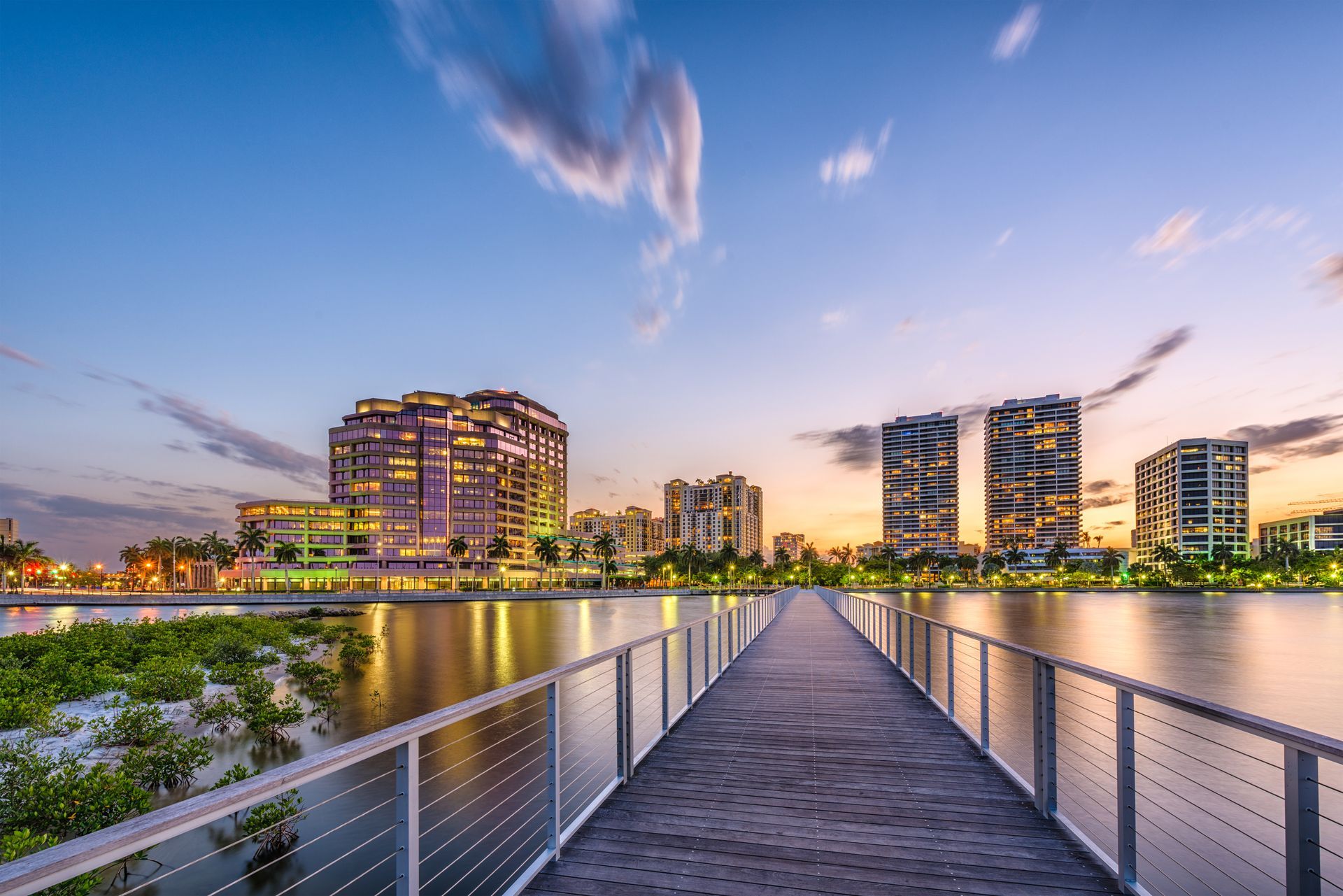 Wooden walkway over water towards a cityscape at dusk; golden lights and blue sky.