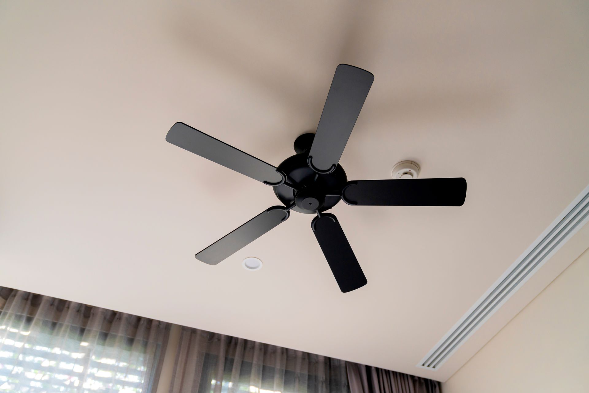 Black ceiling fan on a white ceiling above curtains and a wall air vent