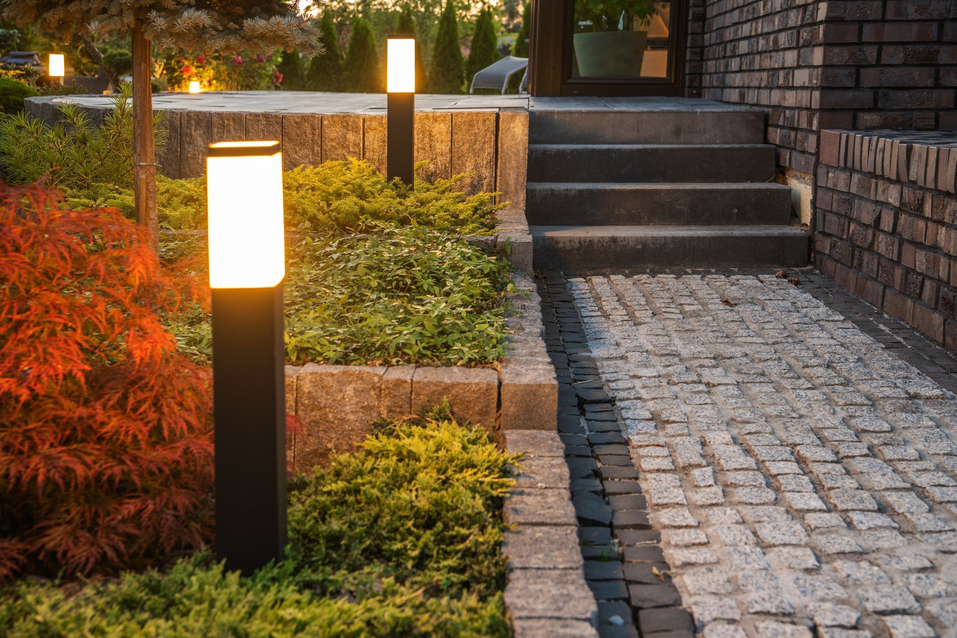 Lit garden path beside brick stairs, with glowing pathway lights and autumn shrubs.