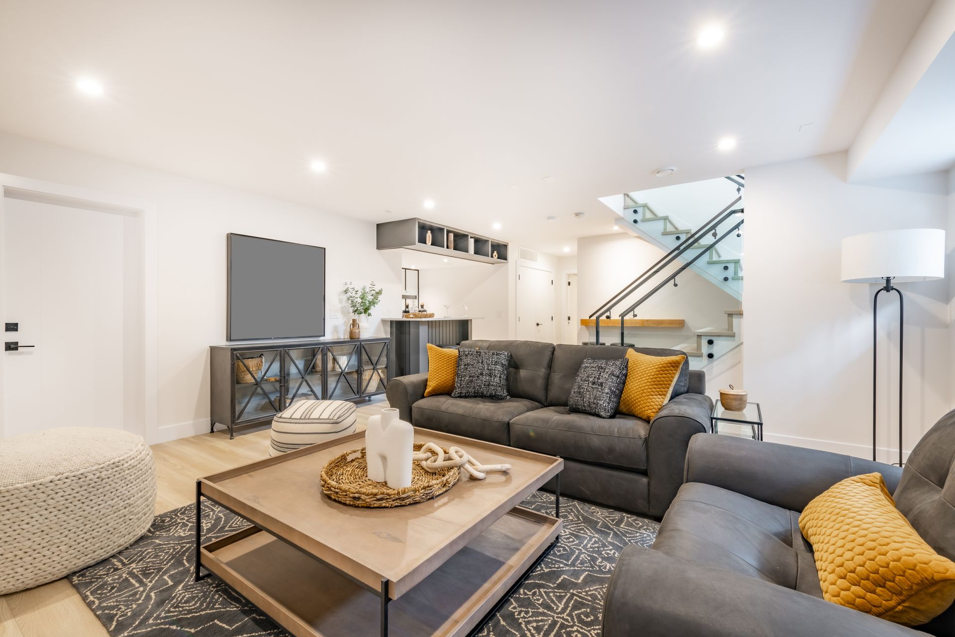Bright modern living room with gray sofas, wood coffee table, TV console, and staircase with glass railing.