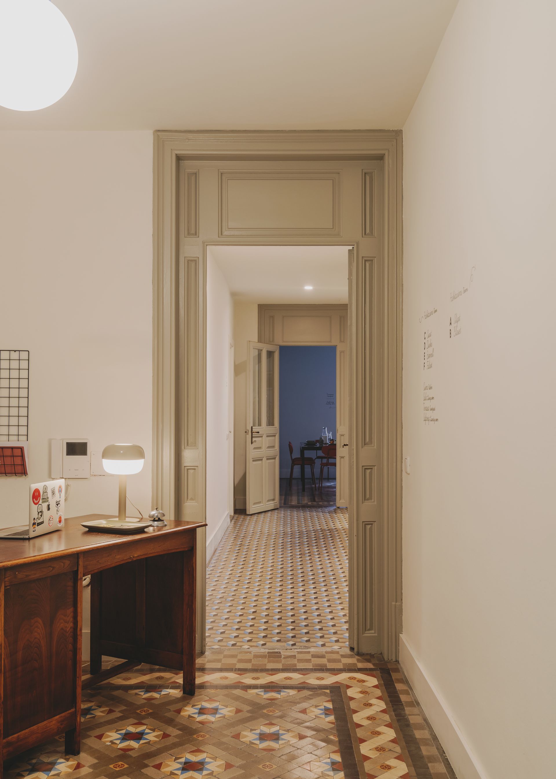 A hallway with a desk and a lamp in it leading to a room.