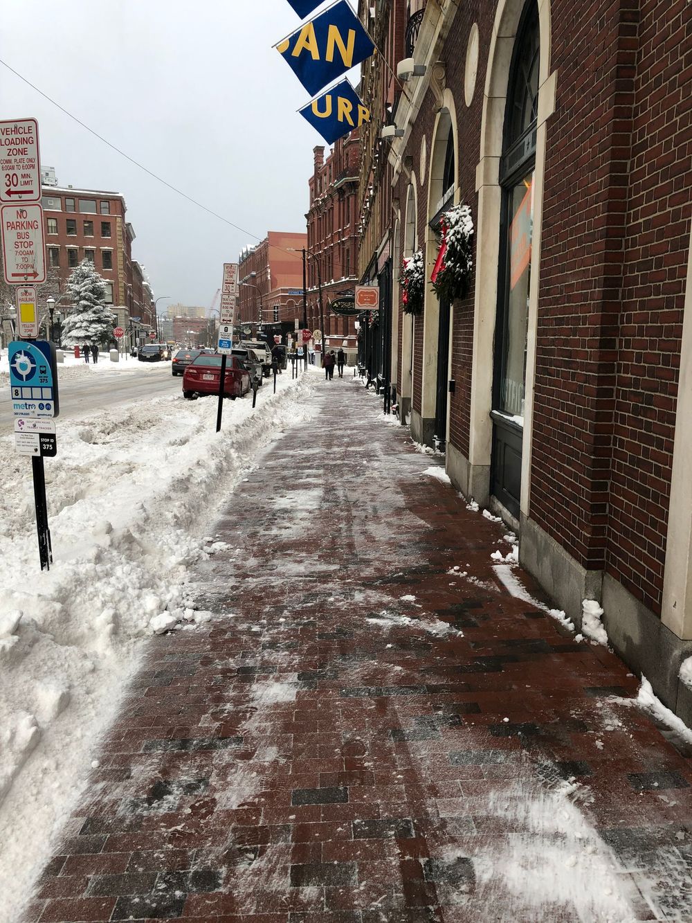 Snowy brick sidewalk next to a red brick building, street lined with snow-covered roads and buildings.