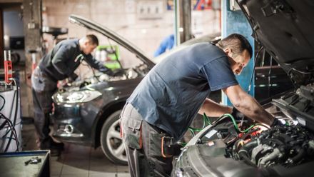Two mechanics working on cars in a garage; one leaning into an engine, the other in the background.