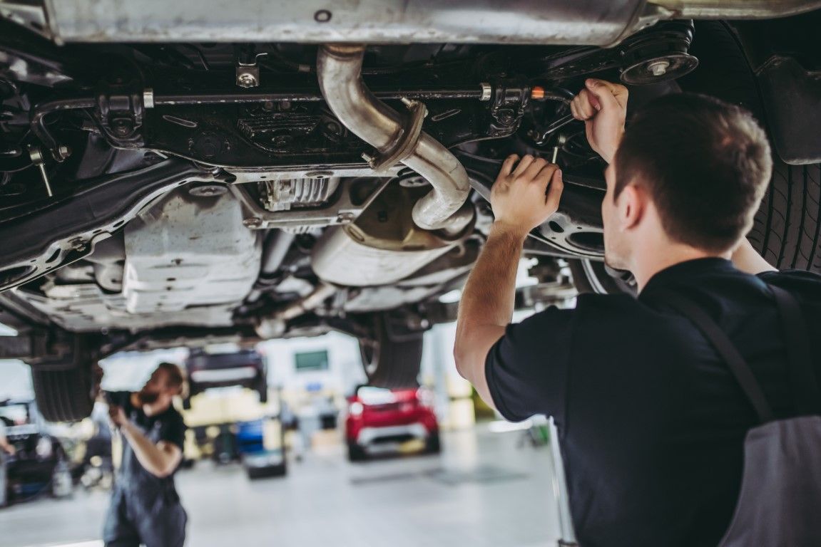 Mechanics working under a car in a garage. One is using tools; the other is in the background.