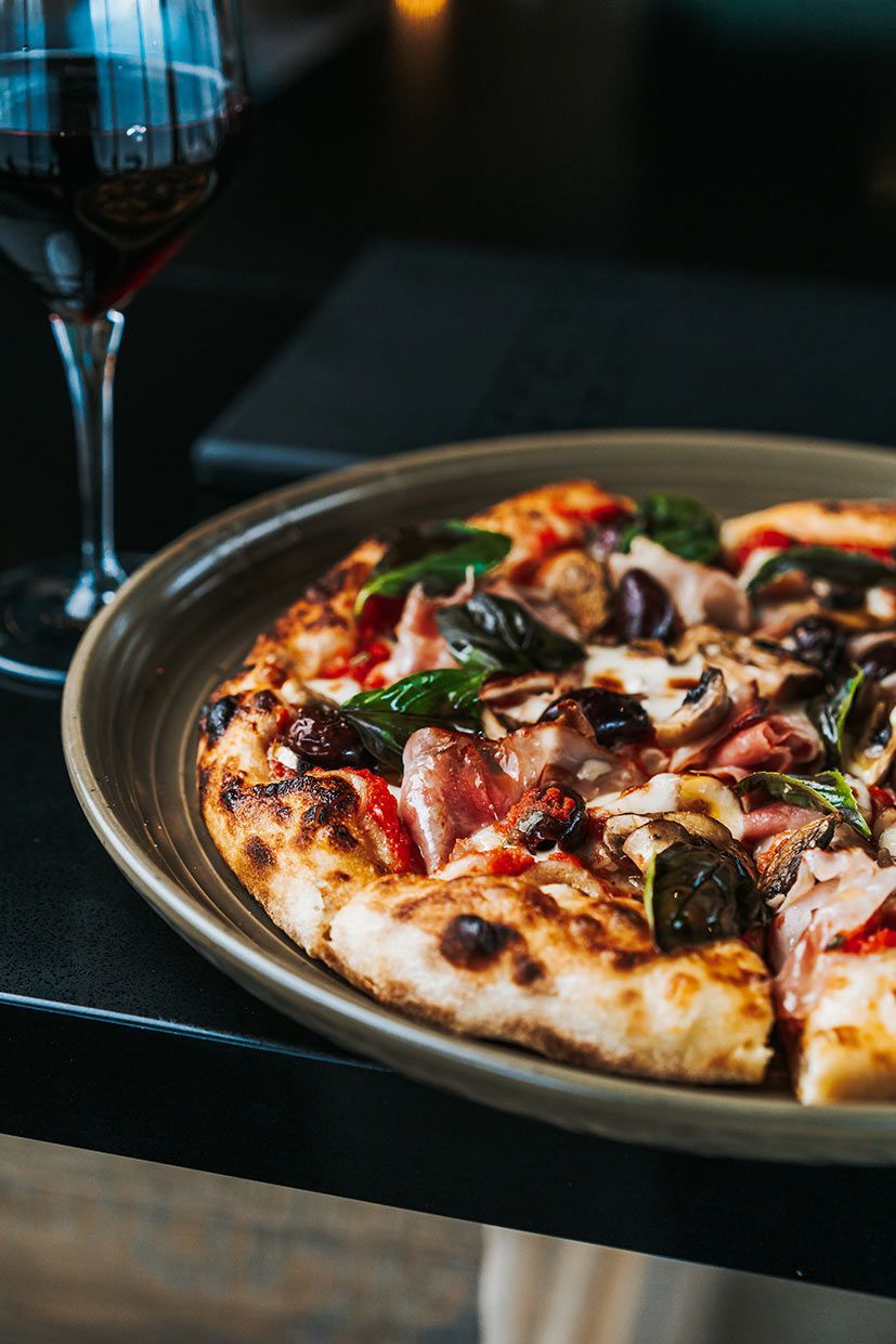 A Pizza on a Plate Next to a Glass of Wine on a Table — Fedeles East Gosford in East Gosford, NSW