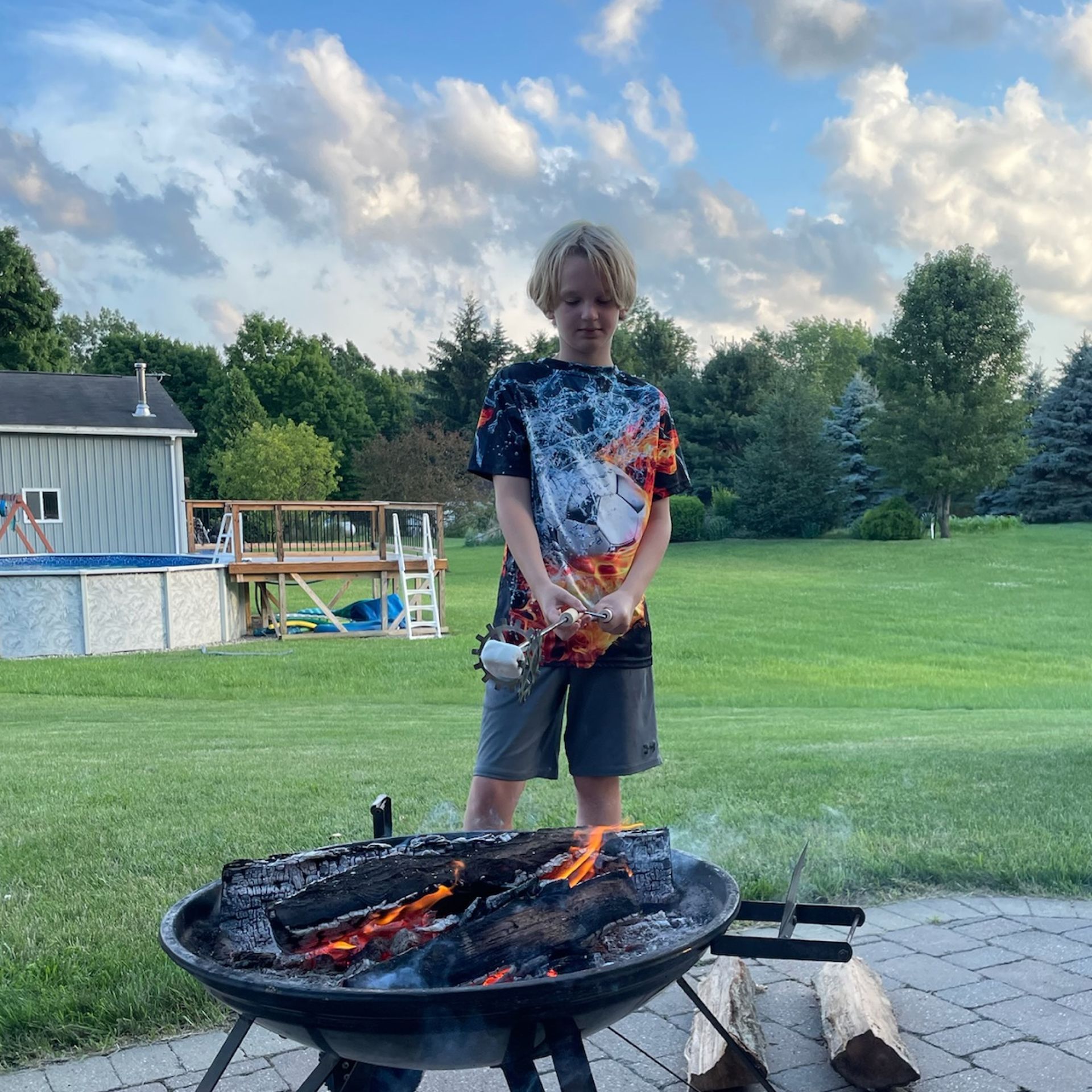 A young boy is standing in front of a grill with a pool in the background.