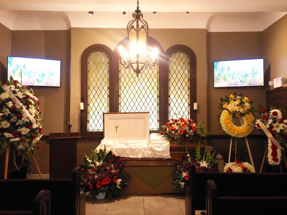 A funeral chapel interior with a white casket, flower arrangements, arched windows, and two screens.