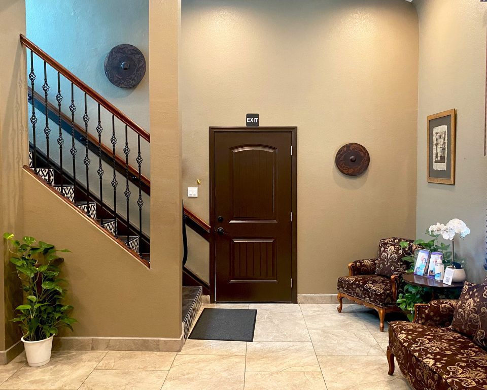 Entryway with stairs, dark brown door, chairs, decorative wall hangings. Neutral wall colors, tiled floor.