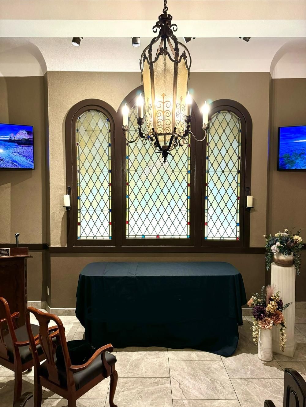 Ceremony space with arched stained-glass windows, dark green table, chandelier, chairs, floral arrangements, and TVs.