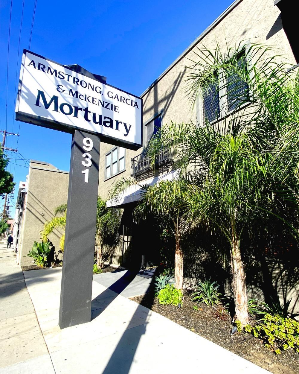 Sign for Armstrong Garcia & McKenzie Mortuary, 931 building, palms in front, gray facade, sunny day.